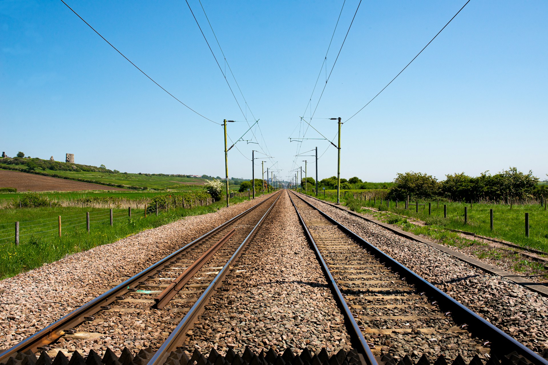 Bahngleise mit Oberleitungsmasten, die durch grüne, hügelige Landschaft unter blauem Himmel führen.
